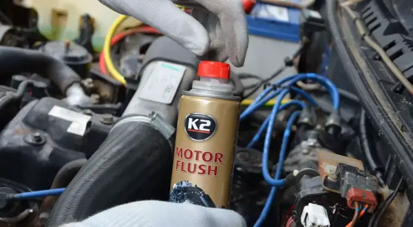 Ręka w rękawiczce trzyma puszkę płukanki do silnika K2 Motor Flush na tle silnika samochodowego.